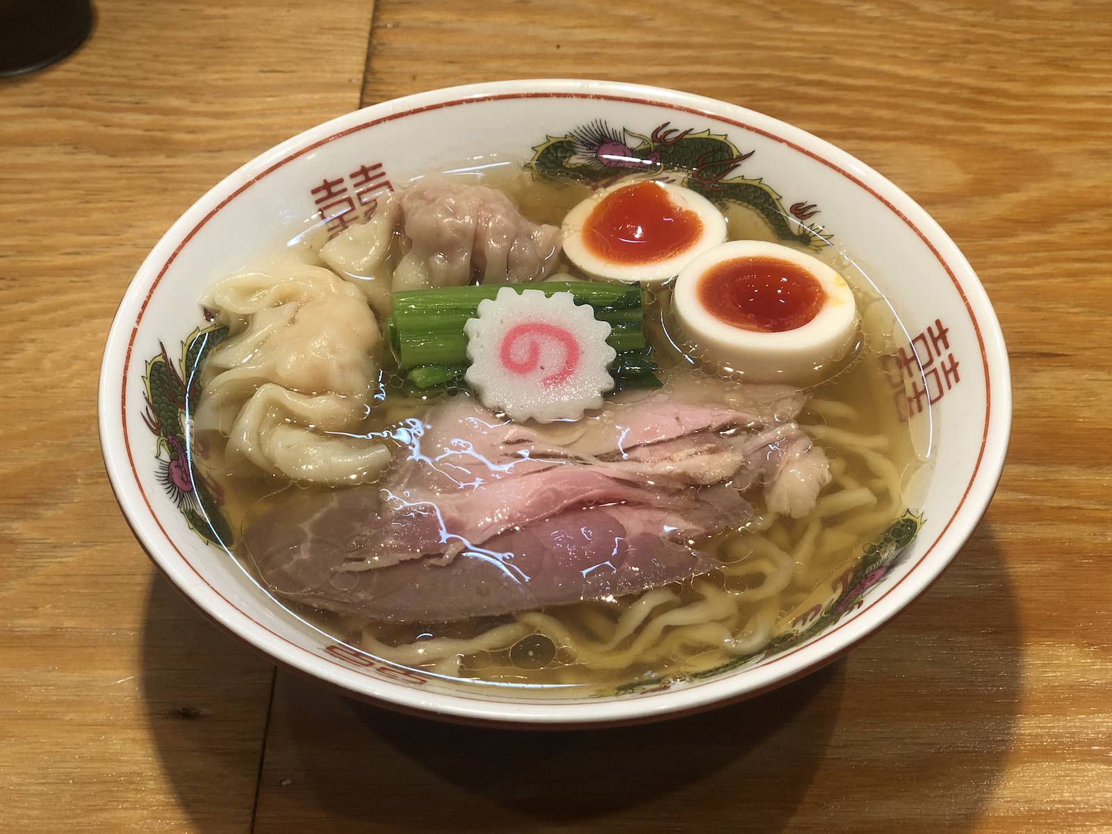 チャーシュー2枚・青菜・半分に切られた味玉・白っぽいワンタン・茶色っぽいワンタン・ナルトが乗った、茶色がかった澄んだスープのラーメン