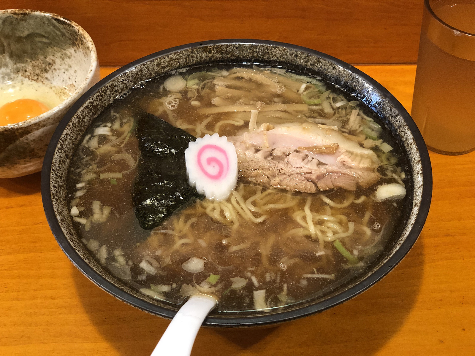 大ぶりの器に並々と注がれた若干濁った濃い目の茶色いスープに輪切りのネギが浮かび、三角形の海苔・崩れそうなバラ肉チャーシュー1枚・なるとが乗った醤油ラーメン。別の器に入った生卵が添えられている。