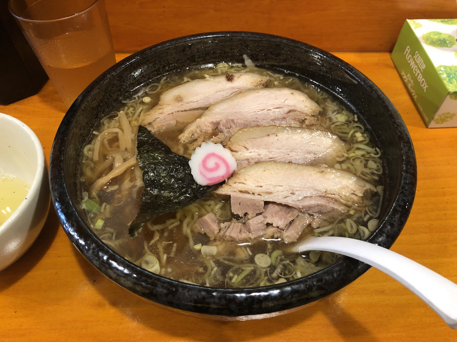 三角形の海苔・崩れかけのバラ肉チャーシュー4枚・なるとが乗り、輪切りのネギが浮かんだ、若干濁った濃い目の茶色いスープの醤油ラーメン。生卵が入っているであろう器が見切れている。