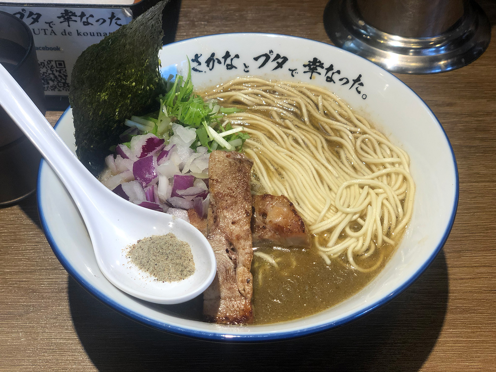 豚ロースステーキのようなものが2切れ・水菜・刻み紫玉葱・海苔が乗った茶濁したスープのラーメン。添えられたレンゲには魚粉が乗っている。