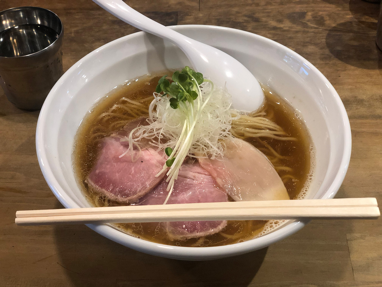 レアな豚チャーシュー2枚・鶏チャーシュー1枚・白髪ねぎネギ・カイワレが乗った、茶色の澄んだスープのラーメン。器の手前よりには割り箸が渡されている。