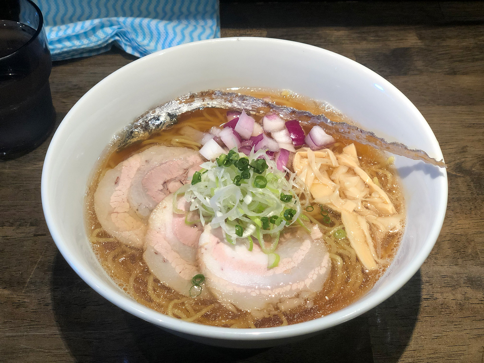 チャーシュー3枚・穂先メンマ・太刀魚の煮干し・刻み紫玉ねぎ・輪切り白ネギ・輪切り青ネギが乗った、茶色い澄んだスープのラーメン