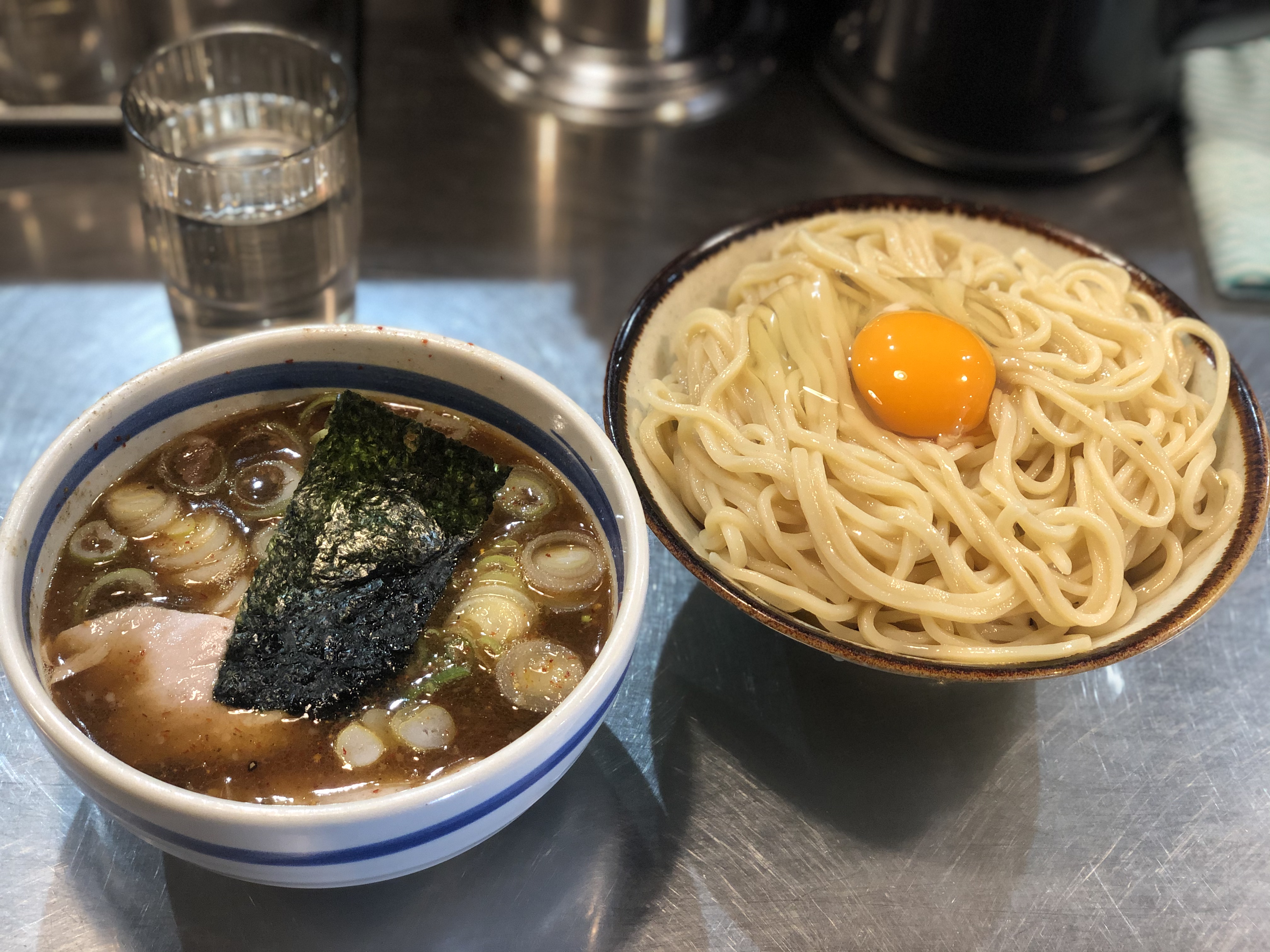 銀色のステンレス風のカウンターに乗ったつけ麺。麺はストレート中太で、茶色い縁取りをされた小ぶりの丼には溢れんばかり盛り付けられ、麺の上には全卵の生卵がのっている。つけ汁は濃い茶色で濁っており、青いラインの入った小ぶりな丼に入っている。スープにはトッピングとしてチャーシューが入っており、海苔1枚、輪切りの白ネギも浮かんでいる。