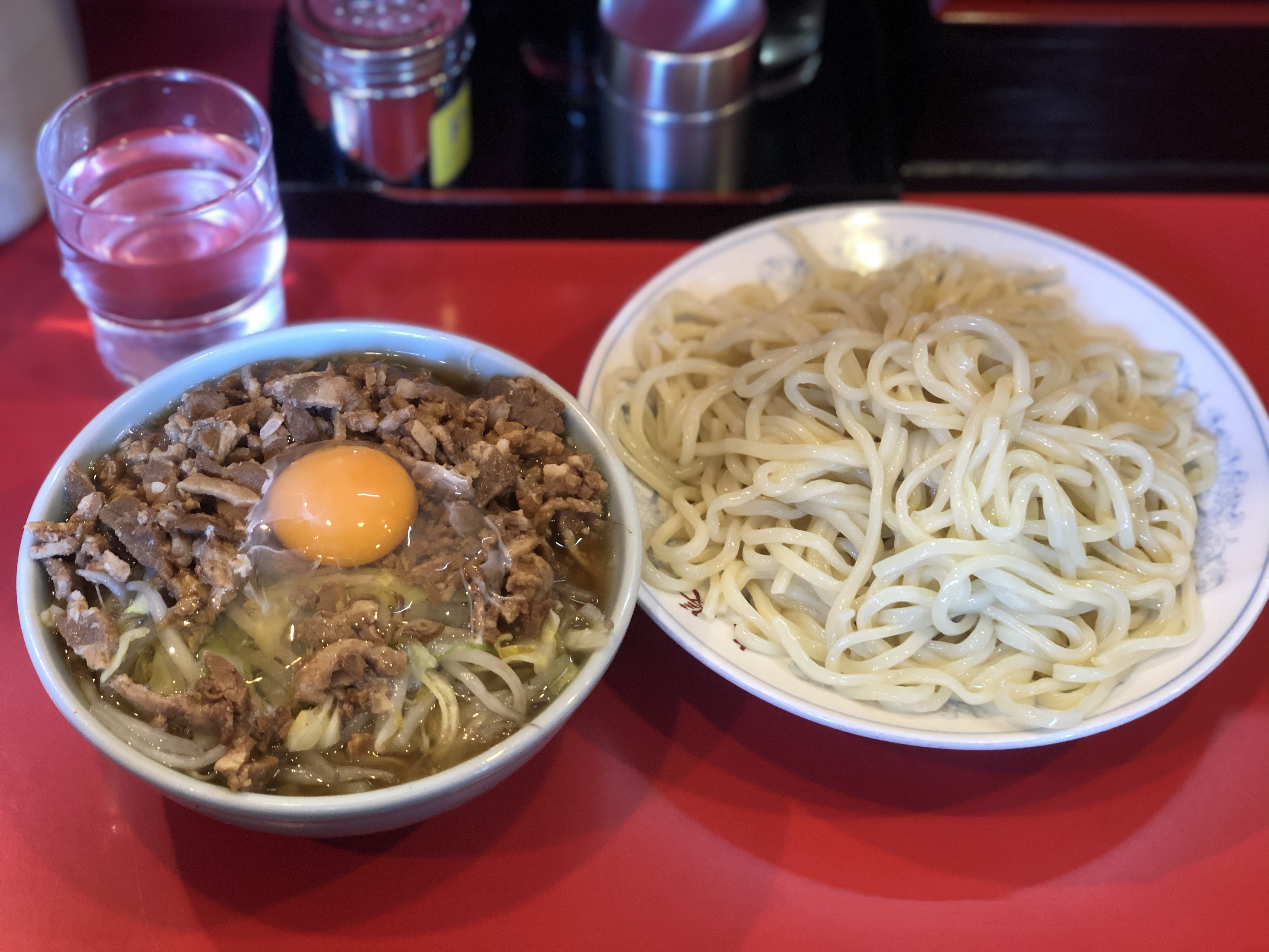 真っ赤なカウンターに乗ったつけ麺。麺は色の淡いストレート中太で、水色絵柄が入った白い皿に盛りつけれている。スープが入っているであろう水色の小鉢には、スープはほぼ見えず細切りにされた大量のチャーシューとキャベツともやしで埋め尽くされており、更にその上には全卵の生卵が乗っている。