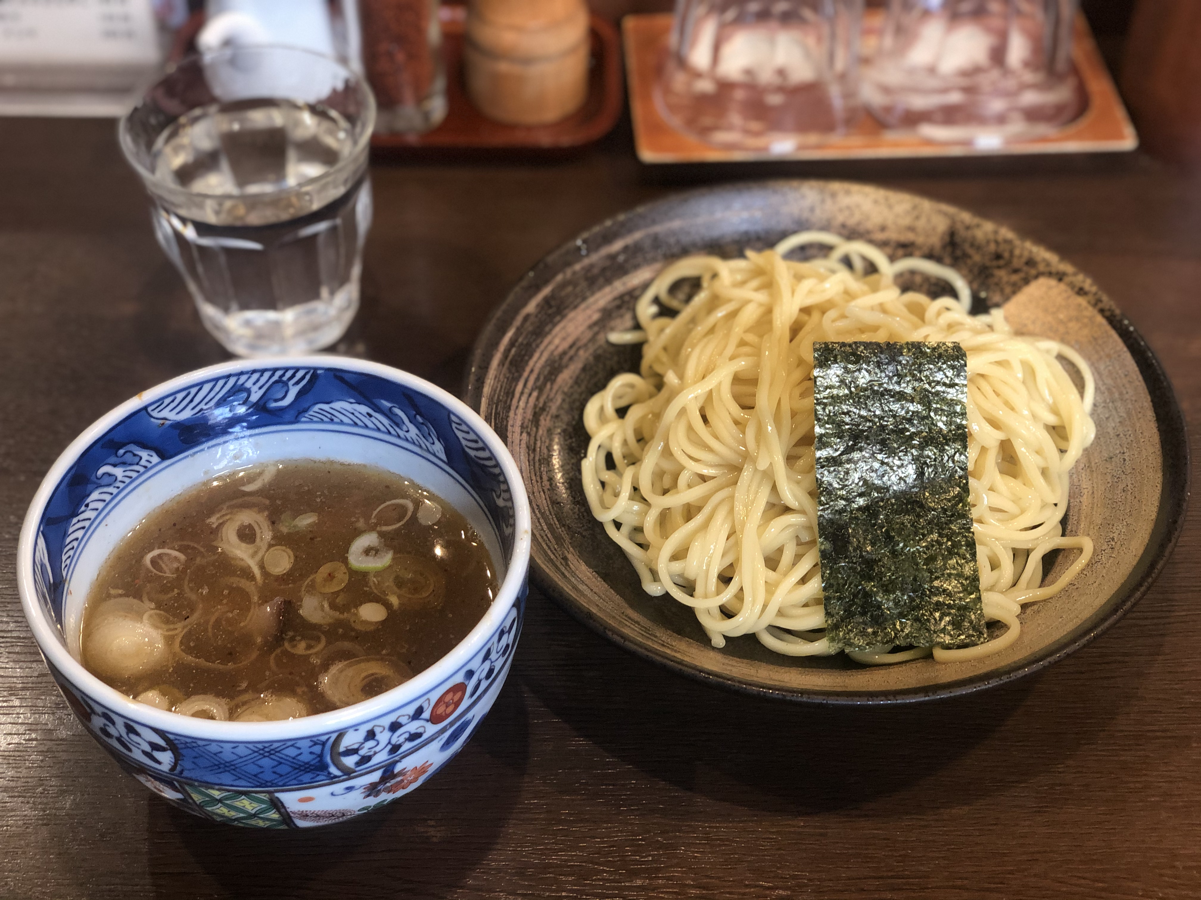 濃い茶色の木目調のカウンターに乗ったつけ麺。麺は中太で、濃い茶色に薄い茶色で模様の入った皿に盛りつけれ、麺の上には海苔が一枚乗っている。つけ汁は濃い色で、青を基調とした模様が描かれたと小鉢に入っており、輪切りの白ネギが浮かんでいる。