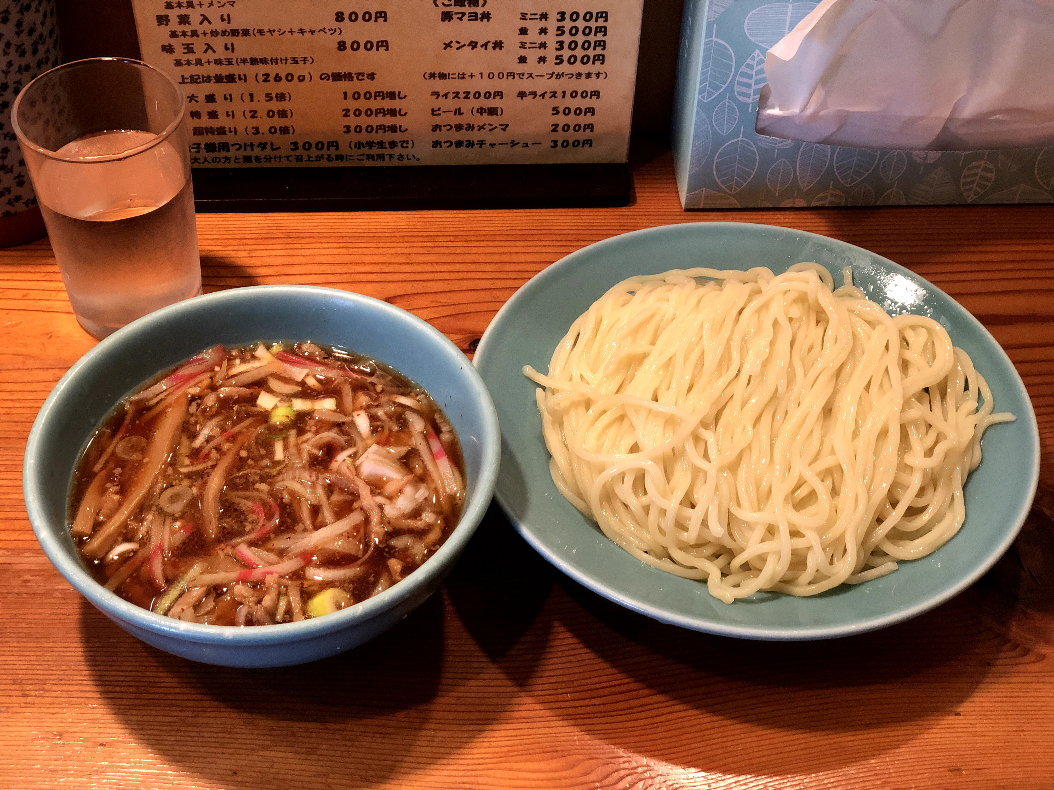 茶色い木目調のカウンターに乗ったつけ麺。麺は色の淡いやや細めの中太で水色の皿に盛り付けられている。つけ汁は濃い茶色で、メンマや細切りにされたかまぼこ・刻んだネギなどが入っている。