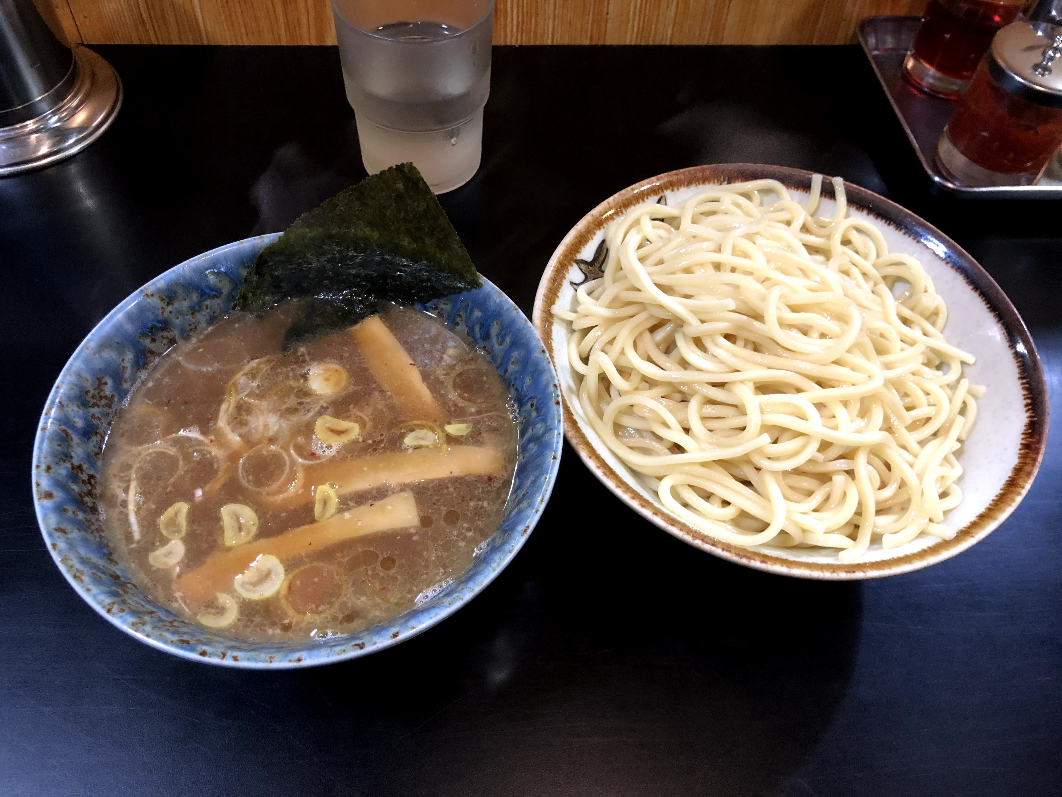 黒いカウンターに乗ったつけ麺。麺は中太で、茶色い縁取りと模様？が入った白っぽい器に盛り付けられている。つけ汁は、少し濁った茶色で、メンマと輪切りの白ネギが入っており、海苔が一枚添えられている。