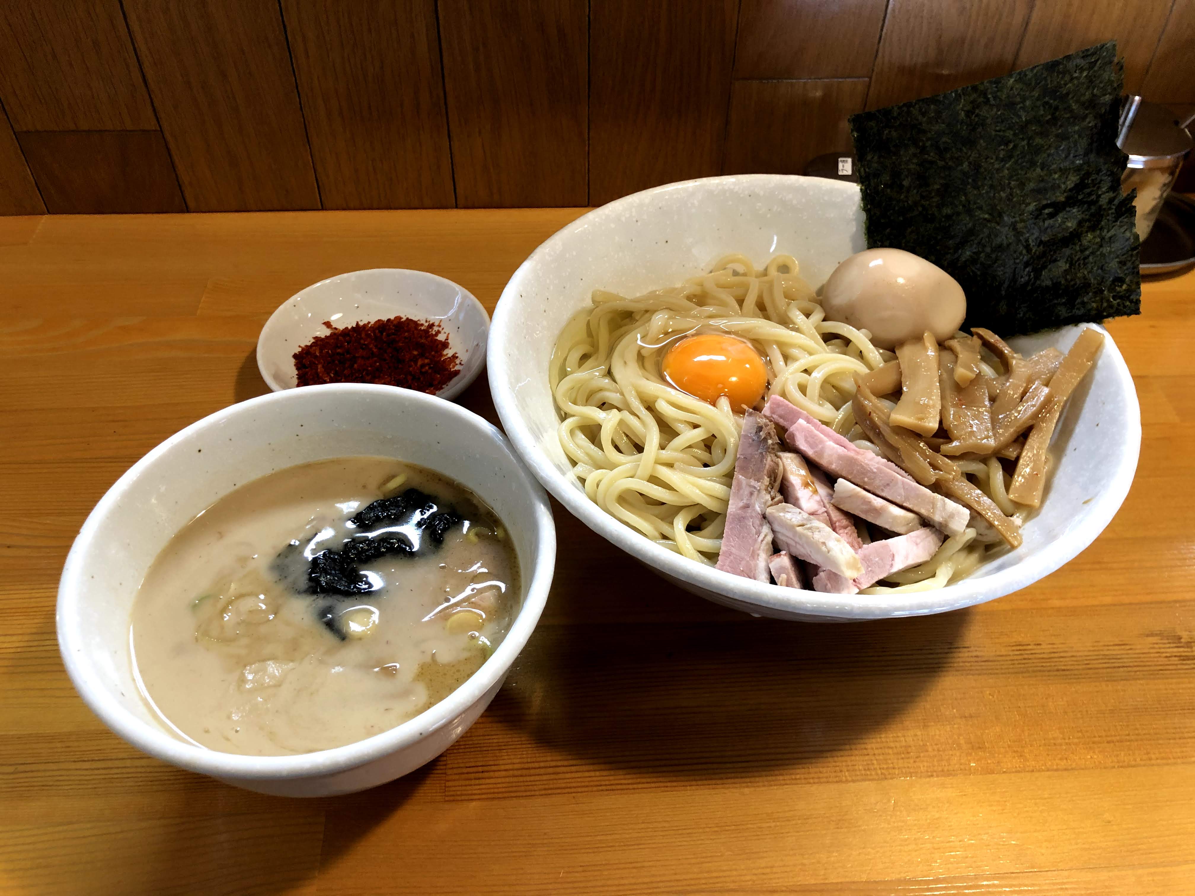 茶色い木目調のカウンターに乗ったつけ麺。麺は太麺で、白い器に盛り付けられ、麺の上には短冊切りになったチャーシューとメンマ・味玉・生玉子・大きめの海苔1枚が乗っている。つけ汁は、白っぽい濁った茶色で、海苔と輪切りの白ネギが入っているように見える。また小さい白い皿に、赤い唐辛子のようなものが添えられている。