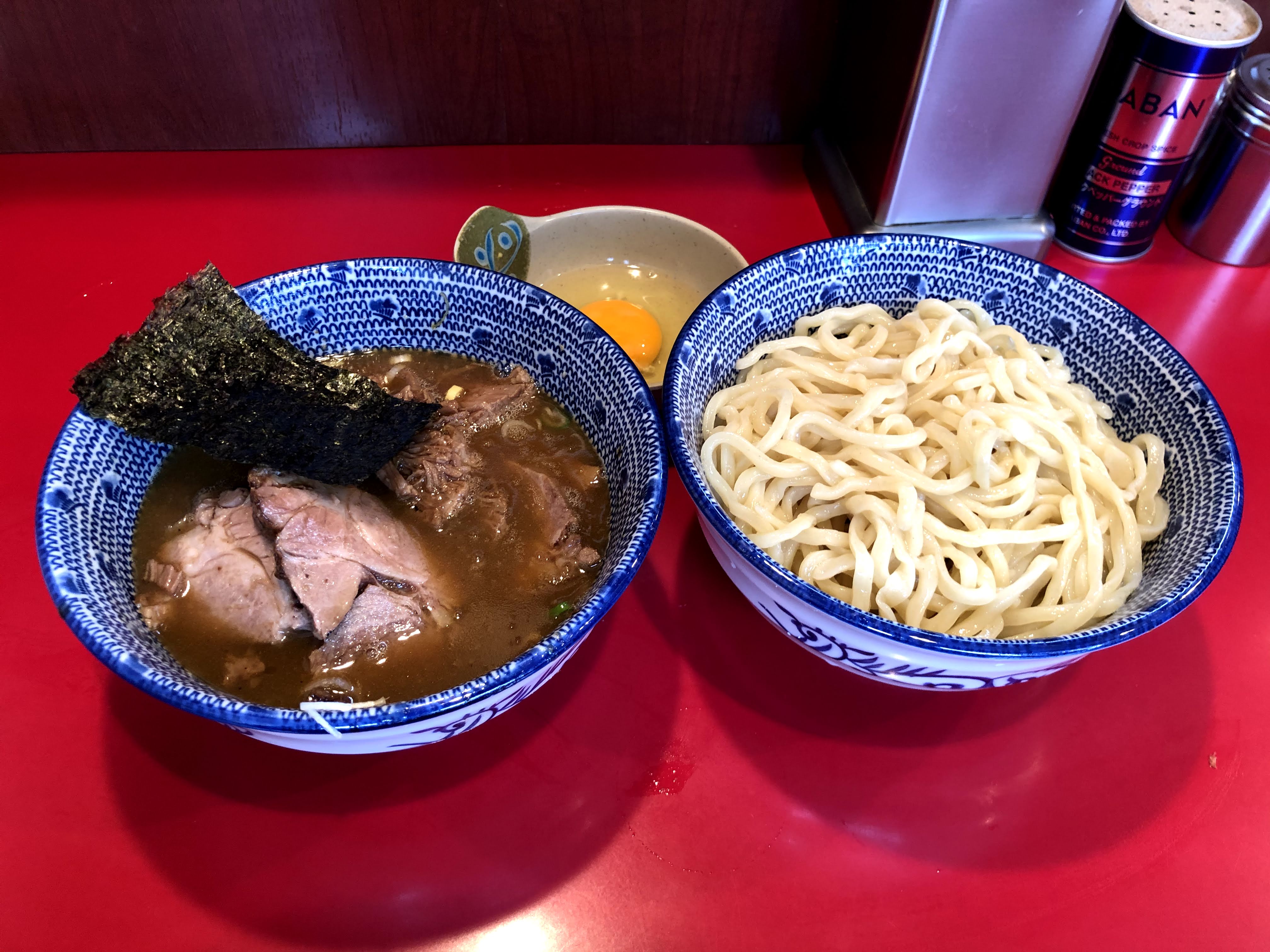 赤いカウンターに乗ったつけ麺。麺は淡い色の太麺で、青い細かい模様が入った器に盛られており、ウェーブがかかっているようにも見える。麺と同じ模様の器に入っているつけ汁は濃い茶色で濁っており、チャーシューが何枚か・海苔・刻んだ白ネギが入っている。また生玉子が入った小さい器も添えられている。