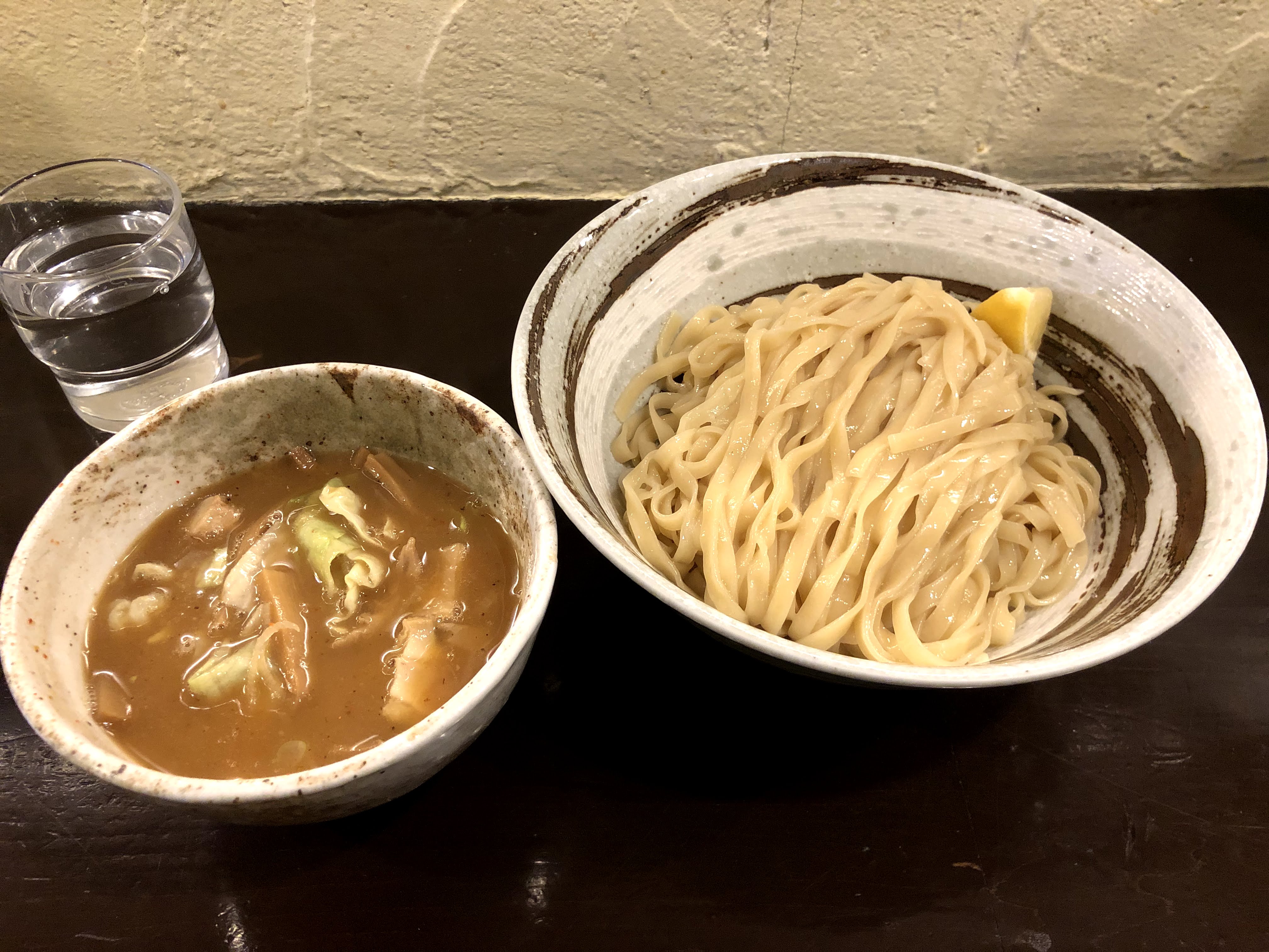 茶色いカウンターに乗ったつけ麺。麺はカップうどんのような？平打ちの麺で、渦を巻いたような模様入った器に盛られており、くし切りを半分にしたレモンが添えられている。ところどころ茶色のシミのような模様の入った白っぽい器に入っているつけ汁は濃い茶色で濁っており、キャベツやメンマが入っている。