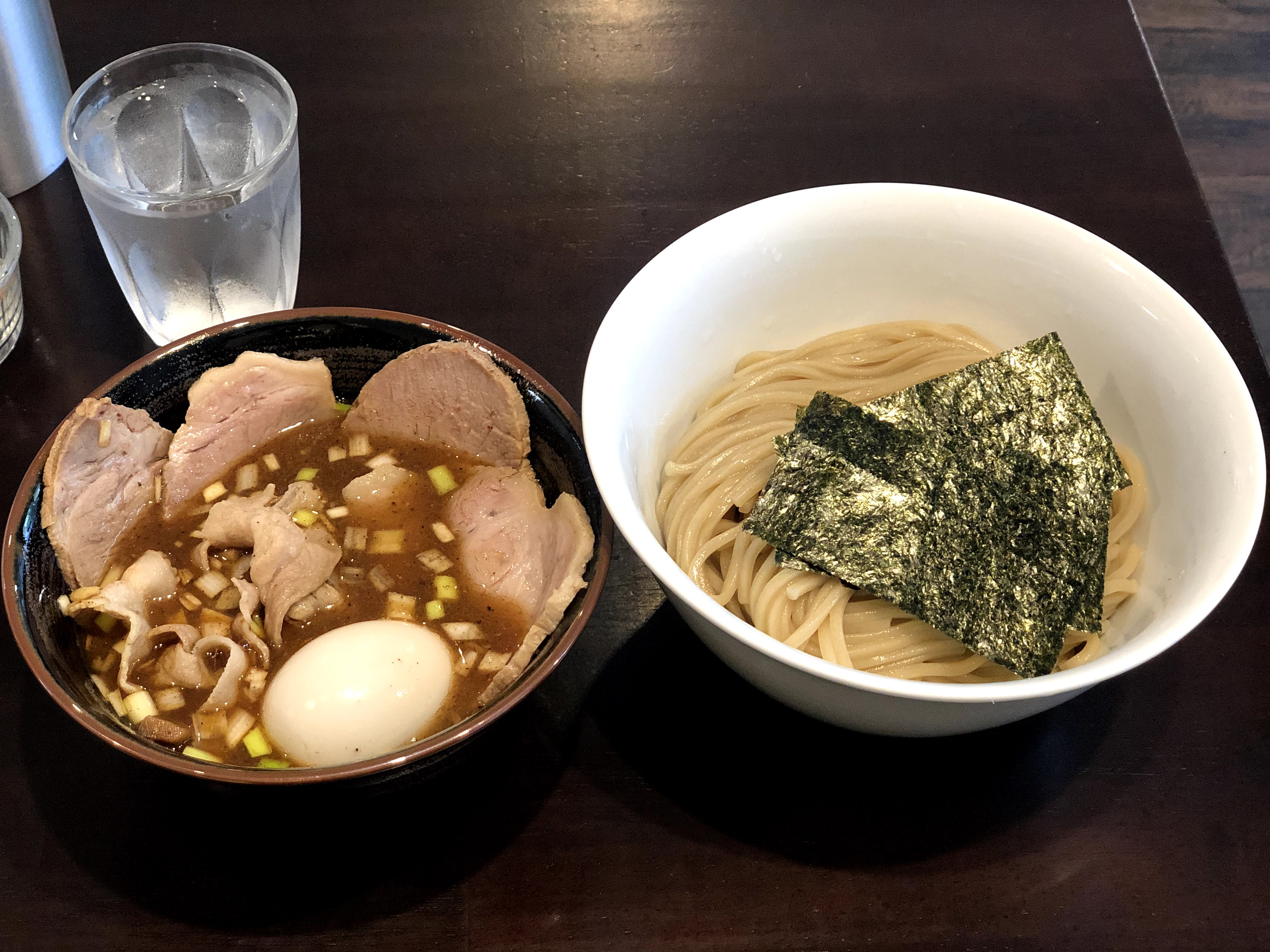 濃い茶色のカウンターに乗ったつけ麺。麺はストレートの太麺で、白い器に盛り付けられており、海苔が3枚乗っている。つけ汁は、濃い茶色で濁っており、茶色で縁取りされた黒い器に入っている。つけ汁には、チャーシュー4枚、薄切りの茹でた豚バラ肉、メンマ・味玉・刻んだ白ネギが入っている。