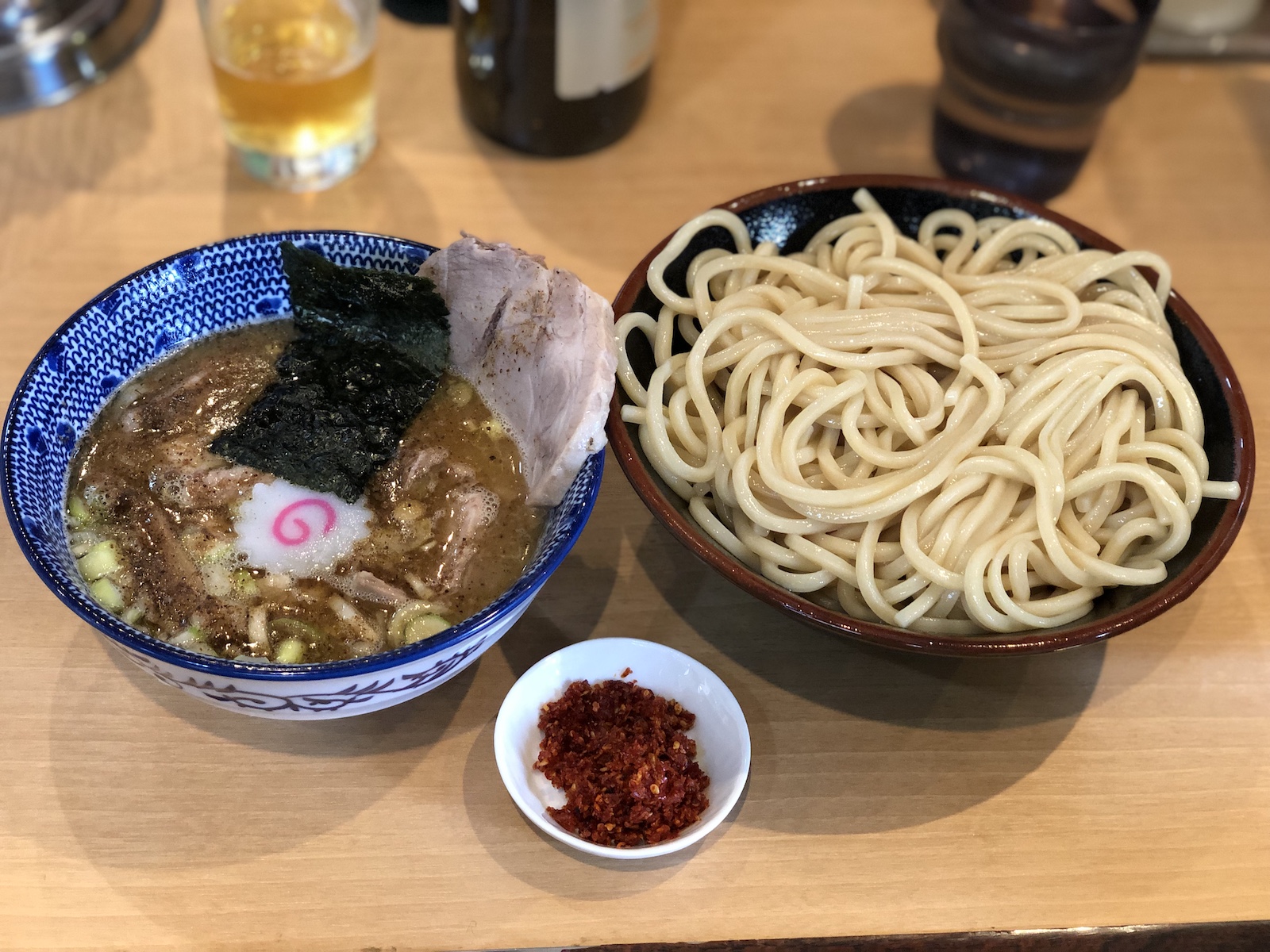白っぽいの木目調のカウンターに乗ったつけ麺。麺は極太で、茶色で縁取りされた黒い器に盛られている。つけ汁は濃い色で、青い模様が入った器に入っており、チャーシュー・海苔・ナルト・小口切りの白ネギが入っていて、角切りチャーシューも沈んでいるようだ。魚粉らしきものがかけられているようにも見える。また小さい白い皿に、赤い唐辛子のようなものが添えられている。